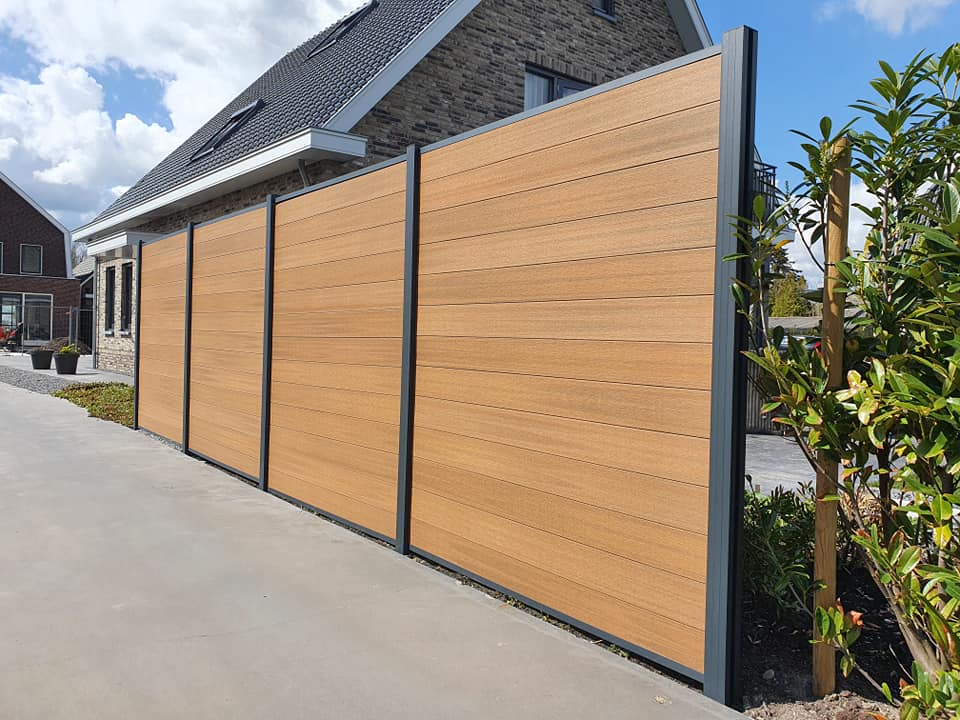 Hoge houtcomposiet schutting in eikenkleur geplaatst als erfafscheiding bij een moderne woning, voorzien van zwarte aluminium profielen